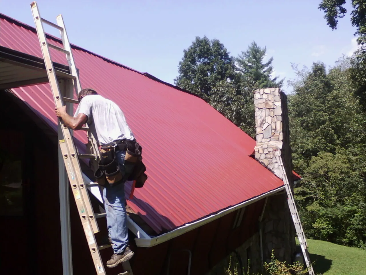 Roofer on a ladder beside a red metal roof and stone chimney — Soffit & Fascia Repair by Roofing Brothers in Califon